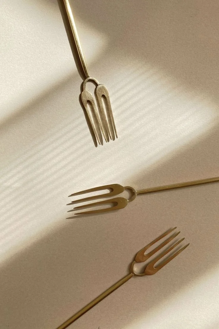 Three gold-colored forks placed on a beige surface with shadows creating horizontal lines.