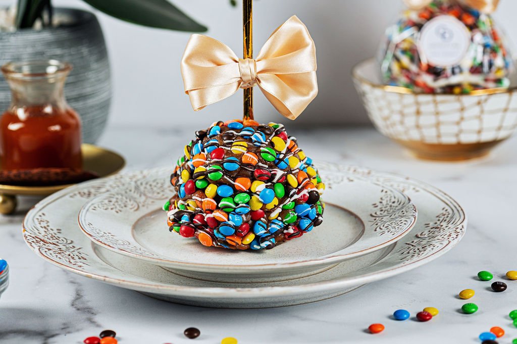 A cake pop coated with colorful candies and chocolate drizzle, topped with a gold bow, on a decorative white plate.