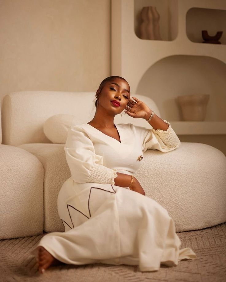 A woman sitting on the floor next to a cream-colored couch, wearing a white dress with beige knitted cuffs, smiling with her eyes closed and resting her head on her hand, in a cozy room with minimalist decor.