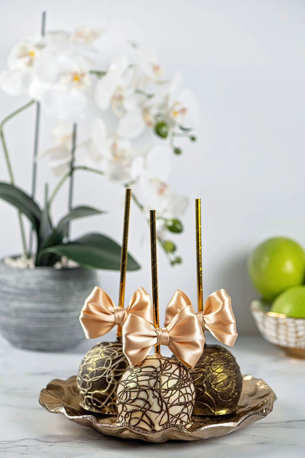 Decorative chocolate-covered treats with gold sticks and satin bows on a silver tray, with white orchids and green apples in the background.