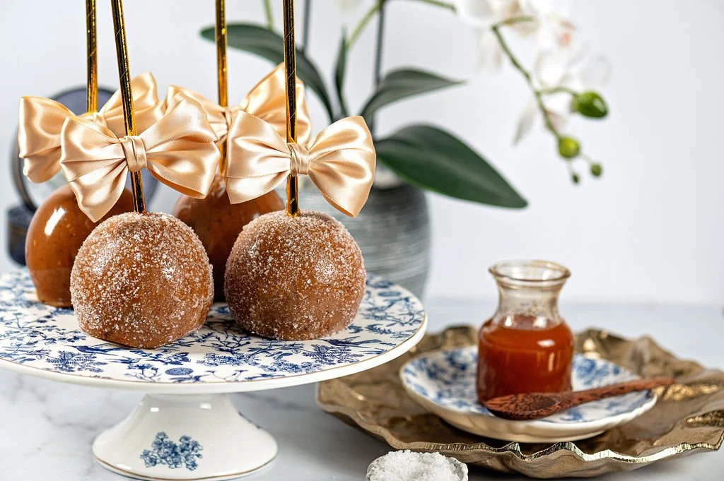 Caramel apples with gold ribbons on a white and blue floral cake stand, a small glass jar of caramel sauce, and a flower arrangement in the background.