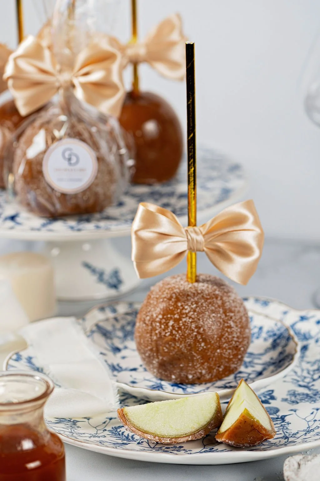 Decorative dessert display with caramel apples covered in sugar, a gold bow, a gold and black straw, and small jars of caramel on a blue and white patterned china.