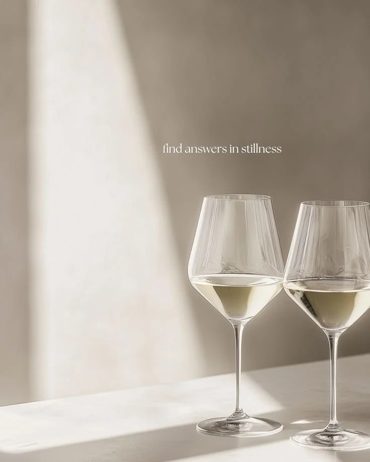 Two wine glasses filled with white wine on a white surface with soft lighting and shadows, with the text 'find answers in stillness' in the background