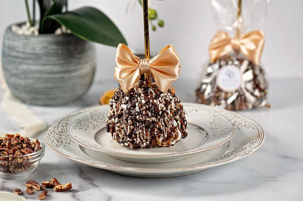 A dessert coated with chopped nuts, drizzled with chocolate, decorated with a satin bow on a stick, placed on a decorative white plate, with a potted plant and another similar wrapped dessert in the background.