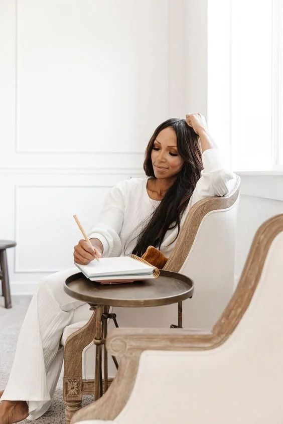 A woman sitting in a beige armchair, writing in a notebook on a small round table, with her left hand resting on her head, in a bright room with white walls and natural light.