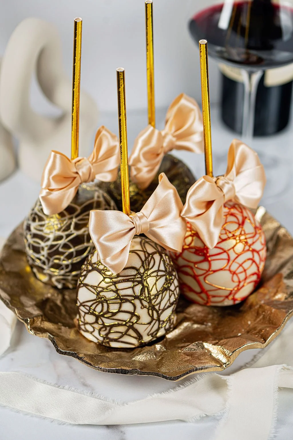 Decorative chocolate cake pops with gold, black, and red designs, each topped with a peach satin bow and a gold stick, arranged on a gold tray.