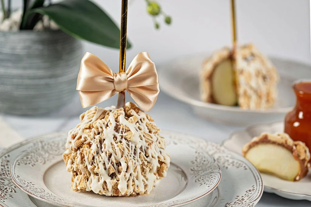 A decorated birthday cake pop topped with a gold bow and white icing, on an ornate white plate. There are other cake pops and desserts in the background.