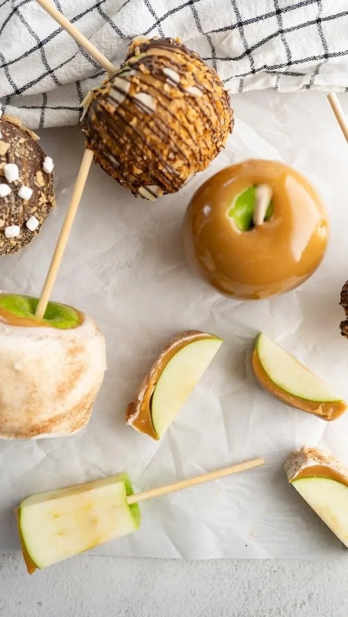 Assorted caramel apples and apples slices with caramel, on white parchment paper.