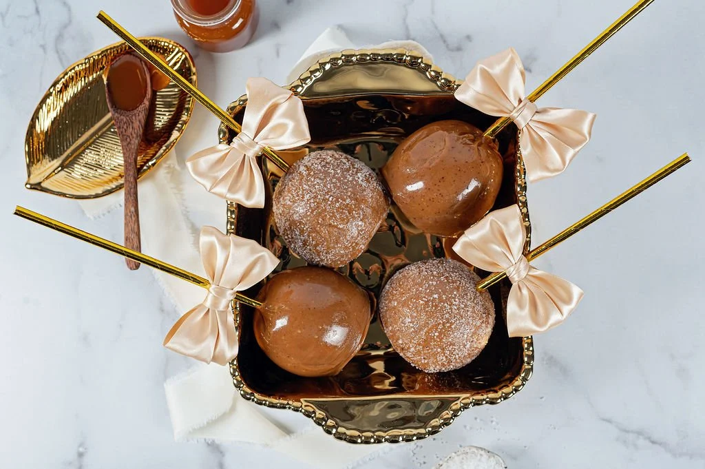 Four caramel-covered cake pops with brown sticks and cream-colored bows in a decorative bowl, with a small bowl of caramel sauce and a jar of caramel in the background on a white marble surface.