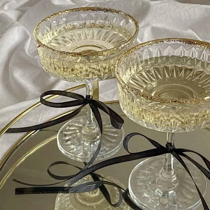 Two elegant glass goblets with gold trim and black ribbons tied around their stems, placed on a mirrored tray with a gold rim, against a white fabric background.