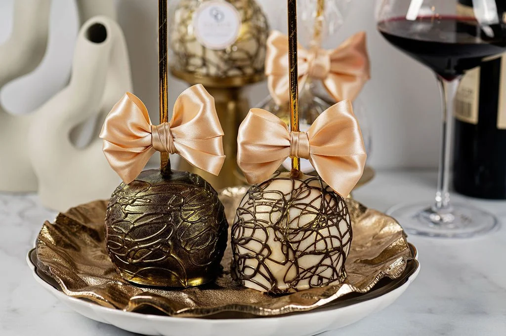 Two decorated chocolates, one with gold drizzles and the other with dark chocolate web pattern, each topped with a peach satin bow, on a gold-edged plate. Glass of red wine and decorative items in the background.