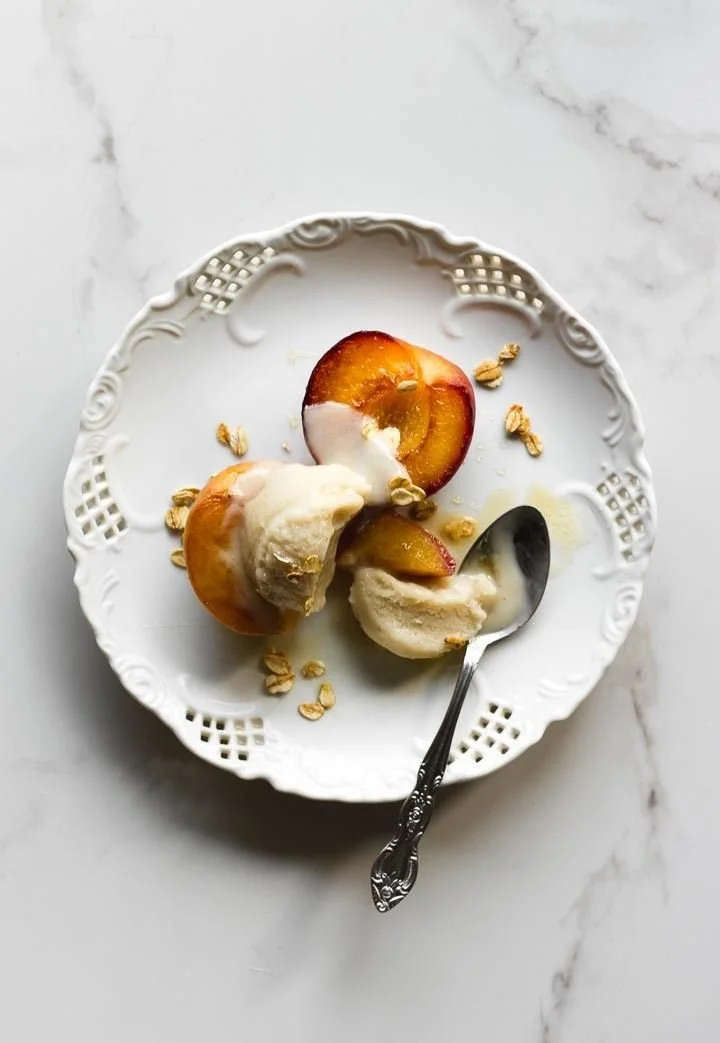 A white ornate plate holds a halved stone fruit with cream and granola, a spoon with a scoop of ice cream.