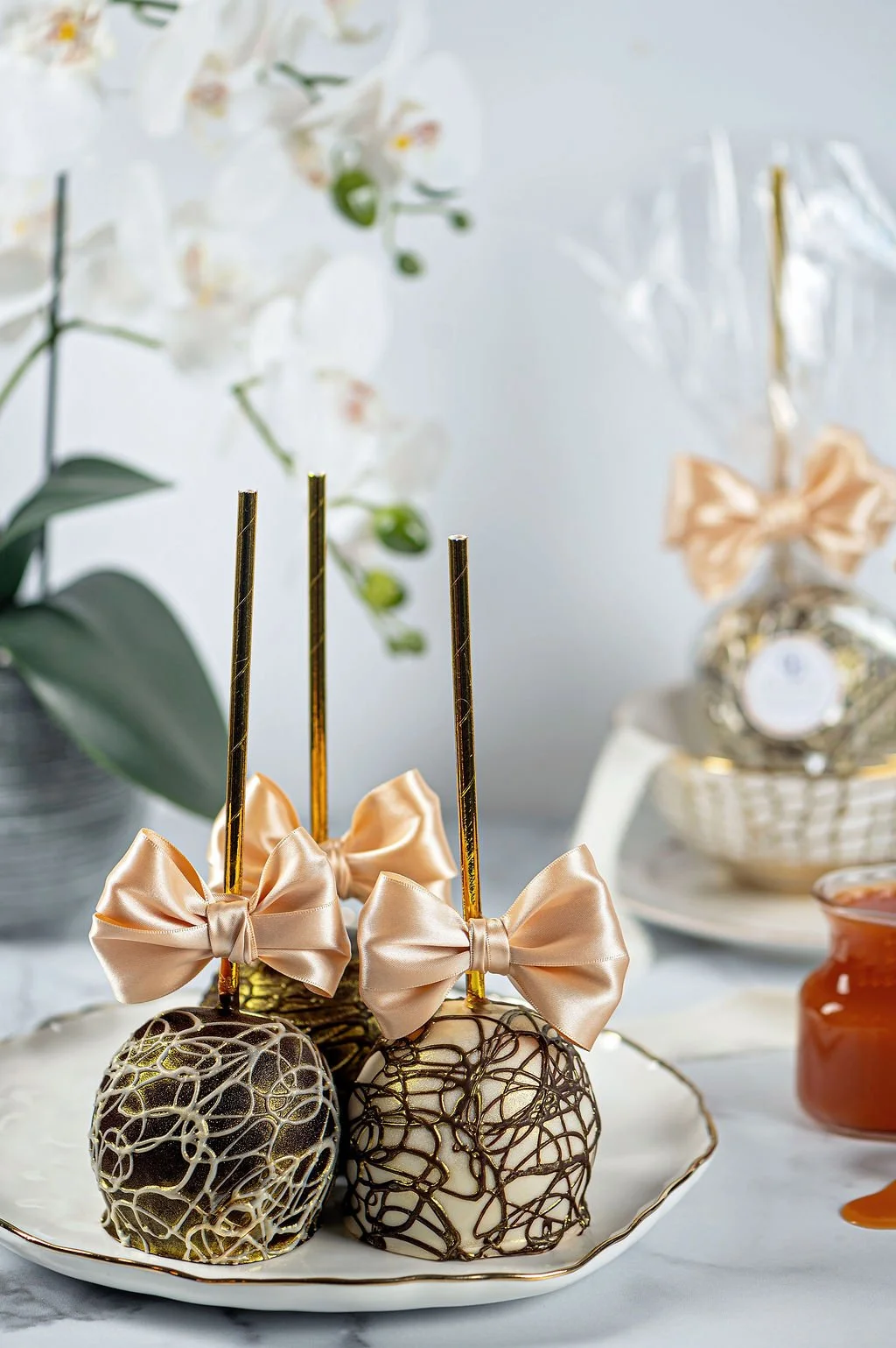 Three decorative chocolate cake pops with gold sticks and peach-colored satin bows on a white plate, with a blurred background of flowers and other desserts.