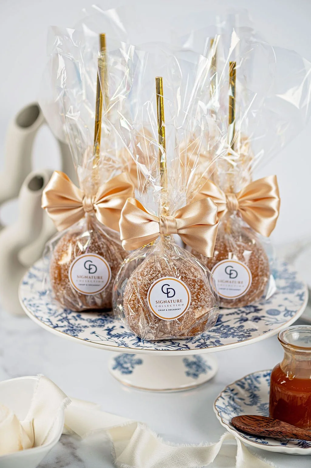 Decorative caramel candies with gold ribbons and twisted sticks, wrapped in clear cellophane, displayed on a blue and white patterned cake stand.