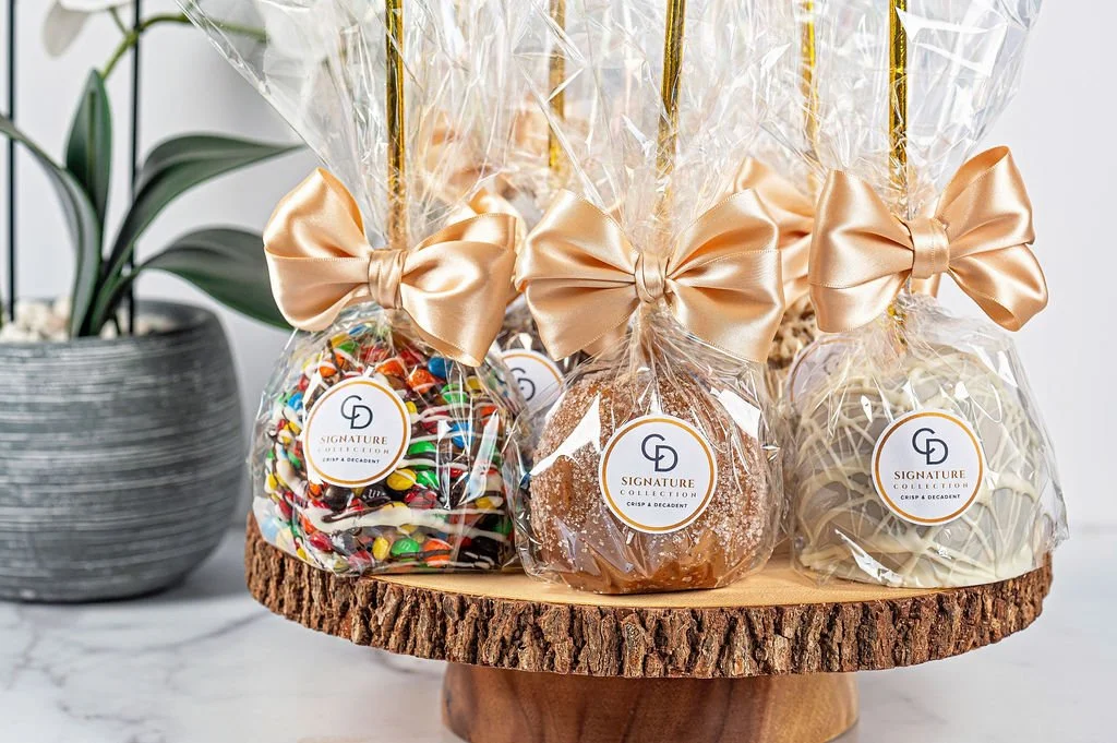 Gift wrapped caramel apple and chocolate lollipop treats with gold bows on a wooden slab, beside a potted plant. 
