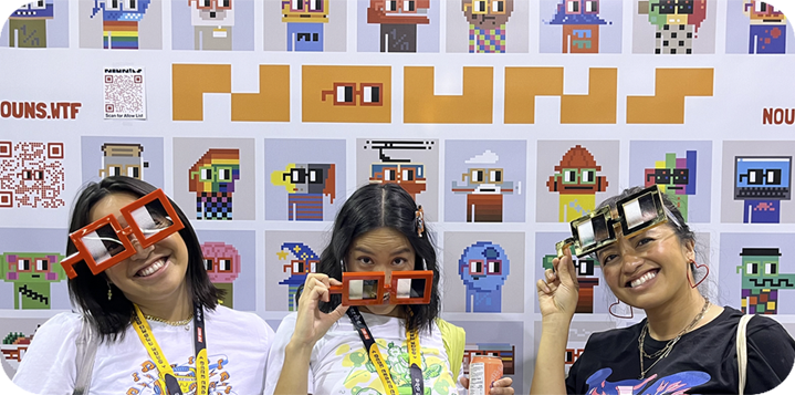 Three women posing in front of a wall covered with colorful pixel art characters, wearing large pixel glasses and smiling.