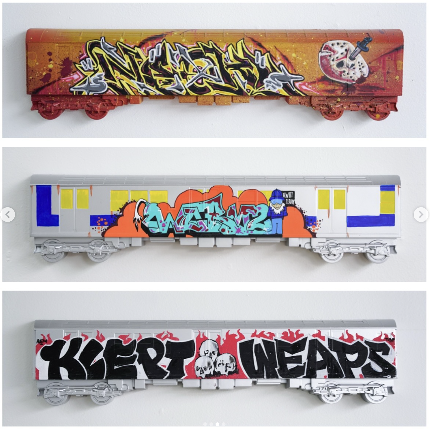 Three model train cars with graffiti art on them, mounted on a wall. The top car has a red background with graffiti text and a paint palette with a brush. The middle car has a silver background with colorful graffiti text, a cartoon character, and bu