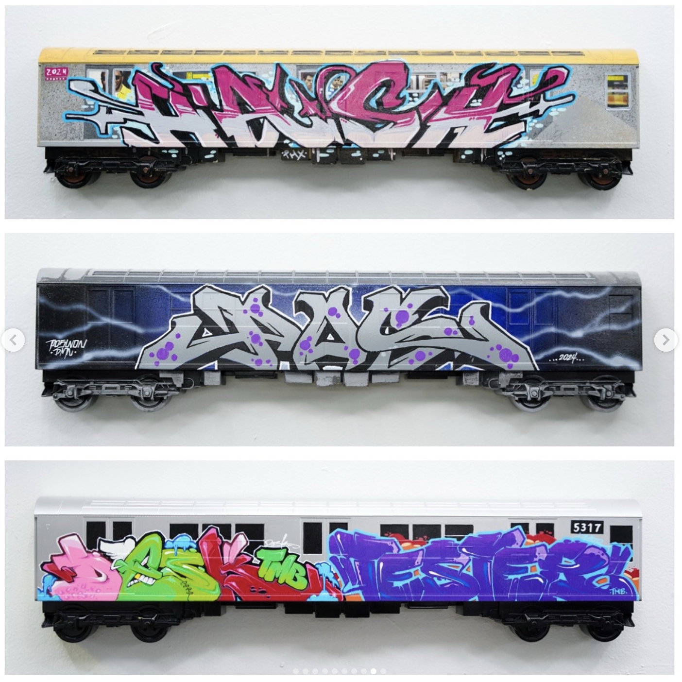 Three model train cars with graffiti art. The top car has pink and blue graffiti with gray background, the middle car has gray graffiti with purple dots and blue background, and the bottom car has multicolored graffiti including pink, green, red, pur