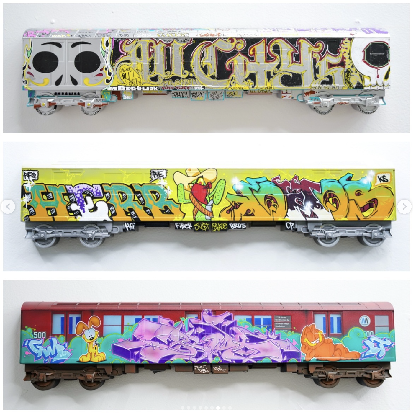 Three model train cars with graffiti art. The top car features black, yellow, and purple graffiti with skull designs. The middle car has yellow, purple, and green graffiti with a cartoon character. The bottom car has a red background with colorful gr