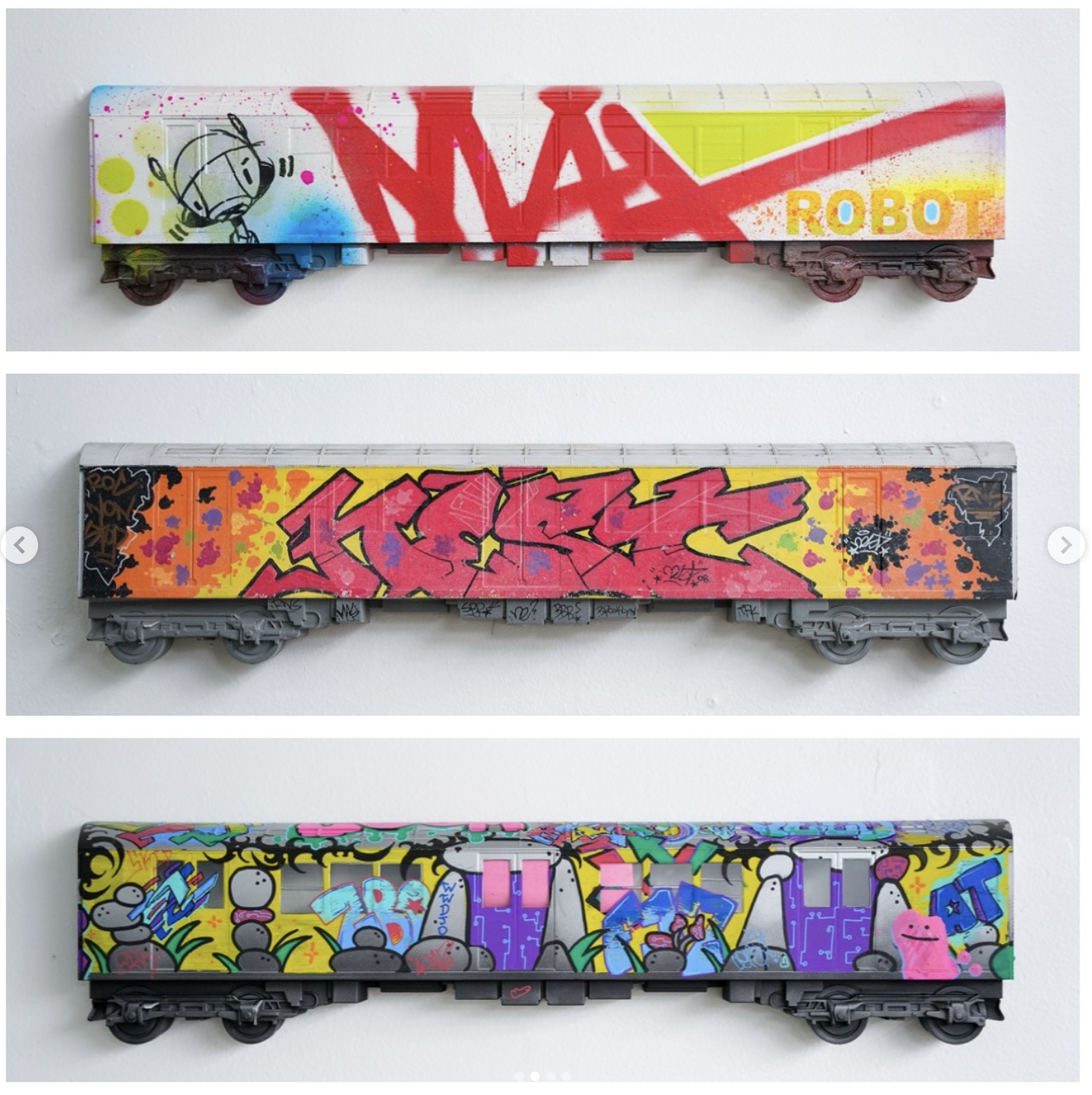 Three model train cars decorated with colorful graffiti art. The top train car has graffiti with the words 'NADA ROBOT' in bold red and yellow. The middle car features graffiti with pink, yellow, and black colors, with abstract designs. The bottom ca