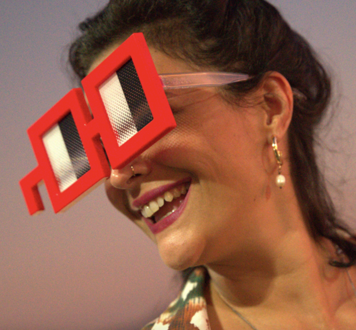 A woman with dark hair and earrings is smiling and wearing large red 3D glasses with black and white lenses, facing to the right.