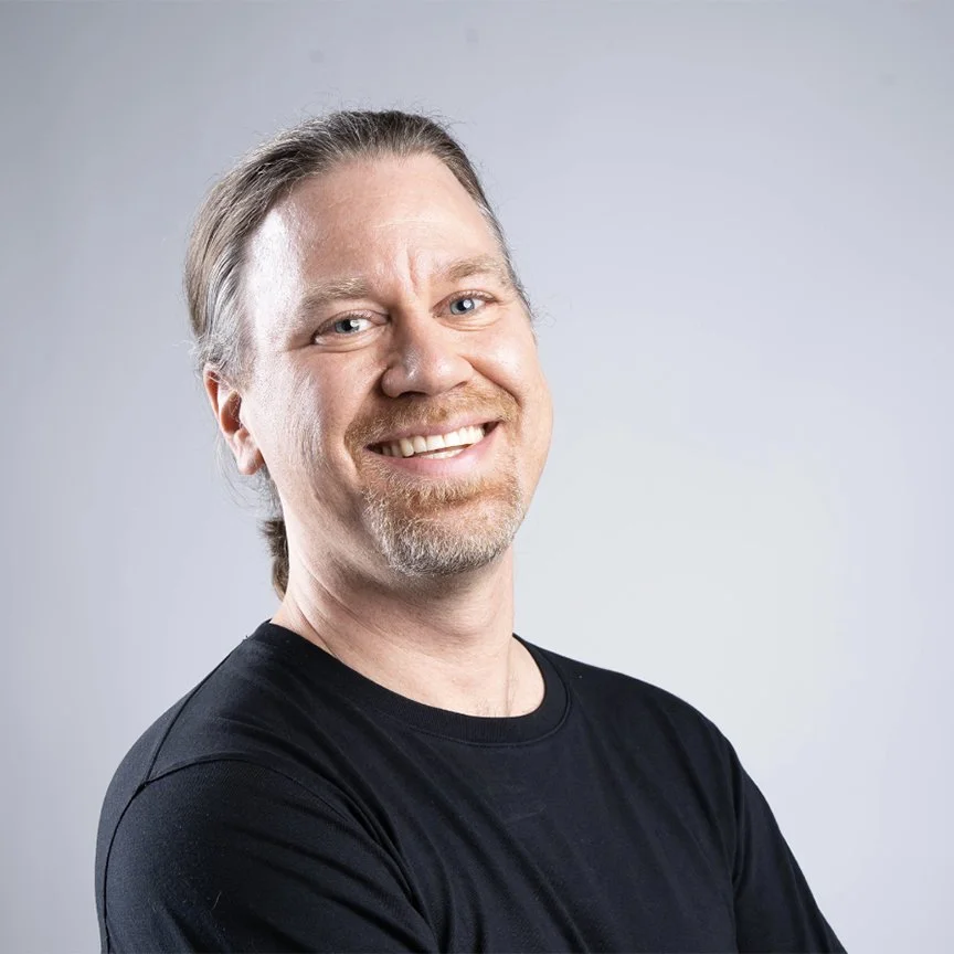 Smiling man with light brown hair and a goatee, wearing a black shirt, against a plain light background.