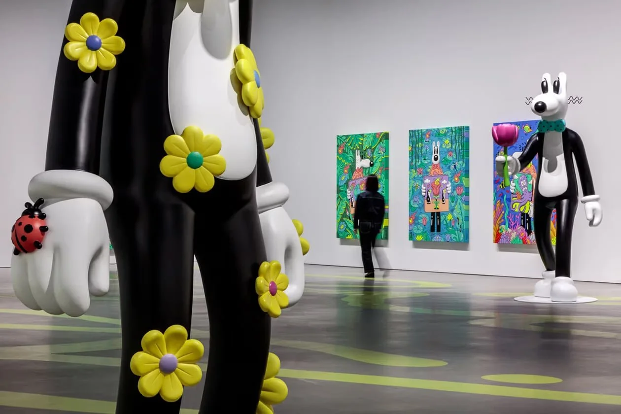Art gallery featuring sculptures of cartoon characters, with one sculpture of a black and white mouse holding a pink flower in the foreground and a large statue of a ladybug-adorned figure, along with vibrant paintings on the wall.