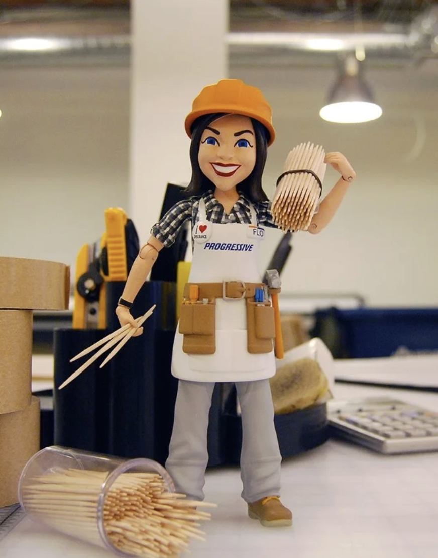 A cartoon doll of a woman in construction worker attire, holding a bundle of matches in her left hand and a small axe in her right hand, standing on a desk with office supplies and tools around.