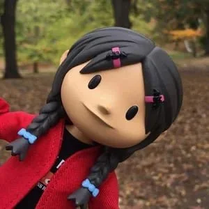 A plush doll of a girl with black braided hair, pink hair clips, and a red jacket, outdoors in a park.