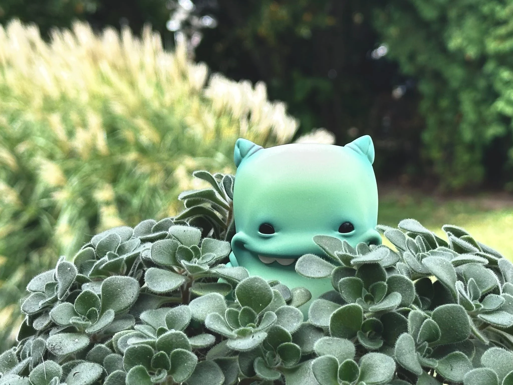 A teal-colored cartoon toy with cat ears and a big smile, positioned among green succulent plants outdoors, with blurred greenery in the background.
