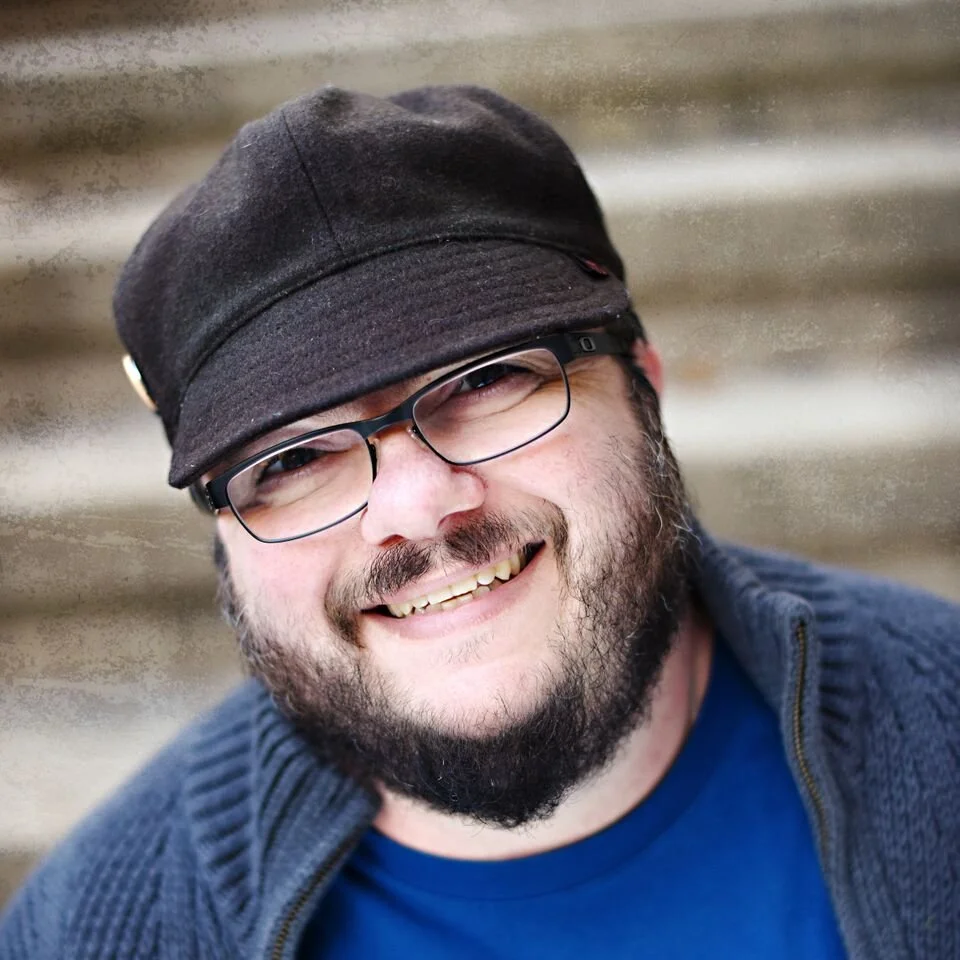 A smiling man with glasses, a beard, wearing a dark cap and a blue shirt.