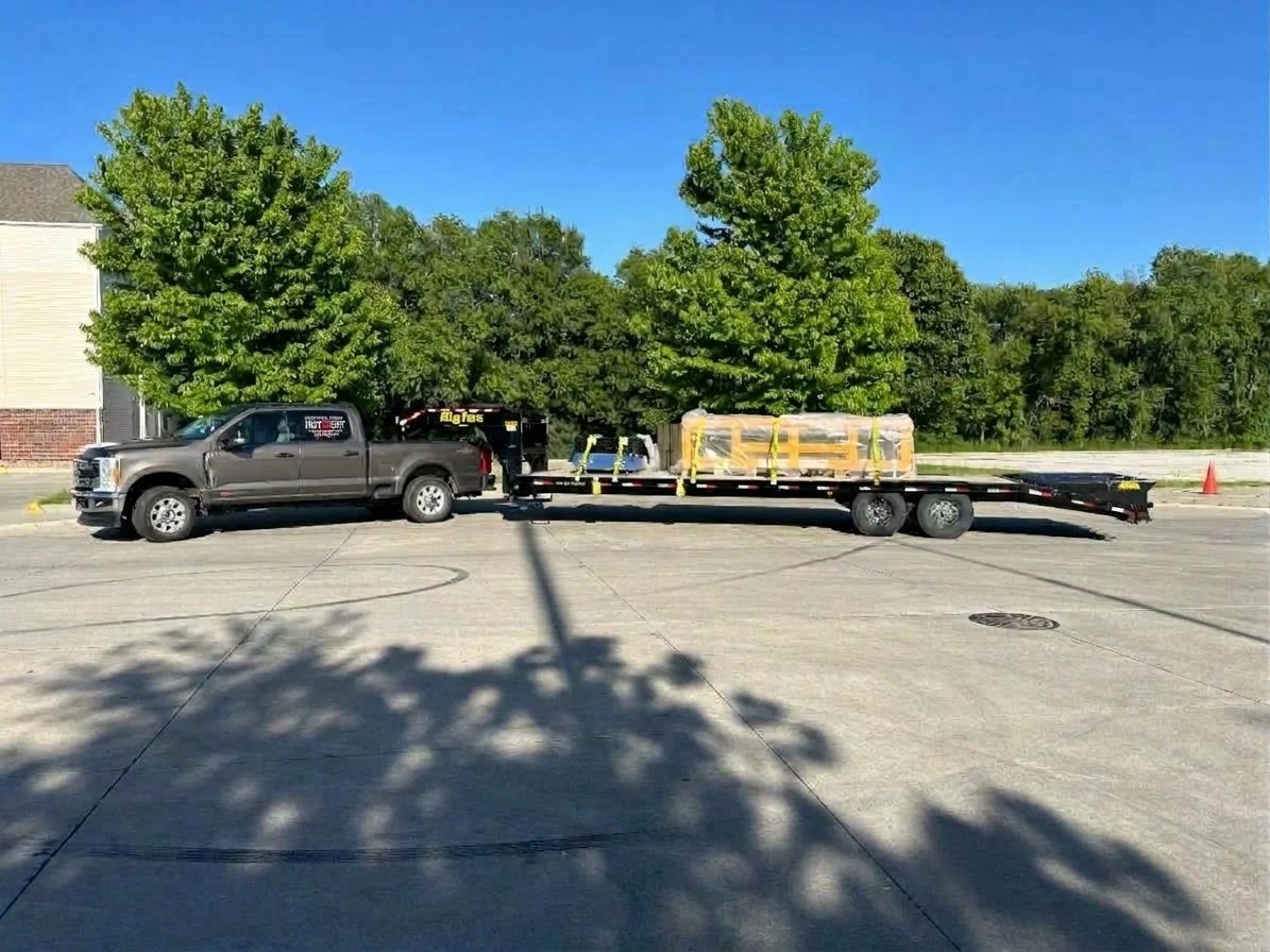 A gray pickup truck towing a flatbed trailer carrying large wrapped packages, parked on a paved area with green trees and a building in the background under a clear blue sky.