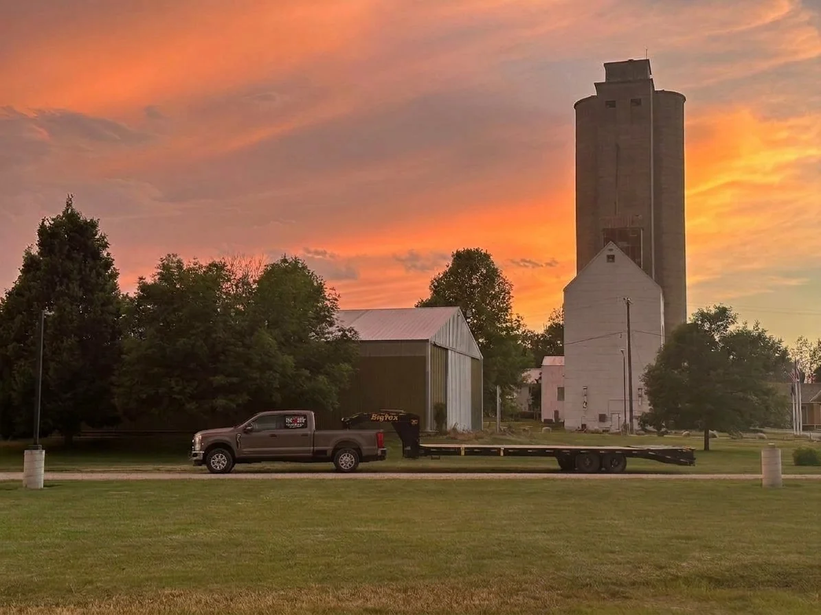 A pickup truck pulling a flatbed trailer parked on a grassy area in front of a barn and silos, with a colorful sunset sky in the background.