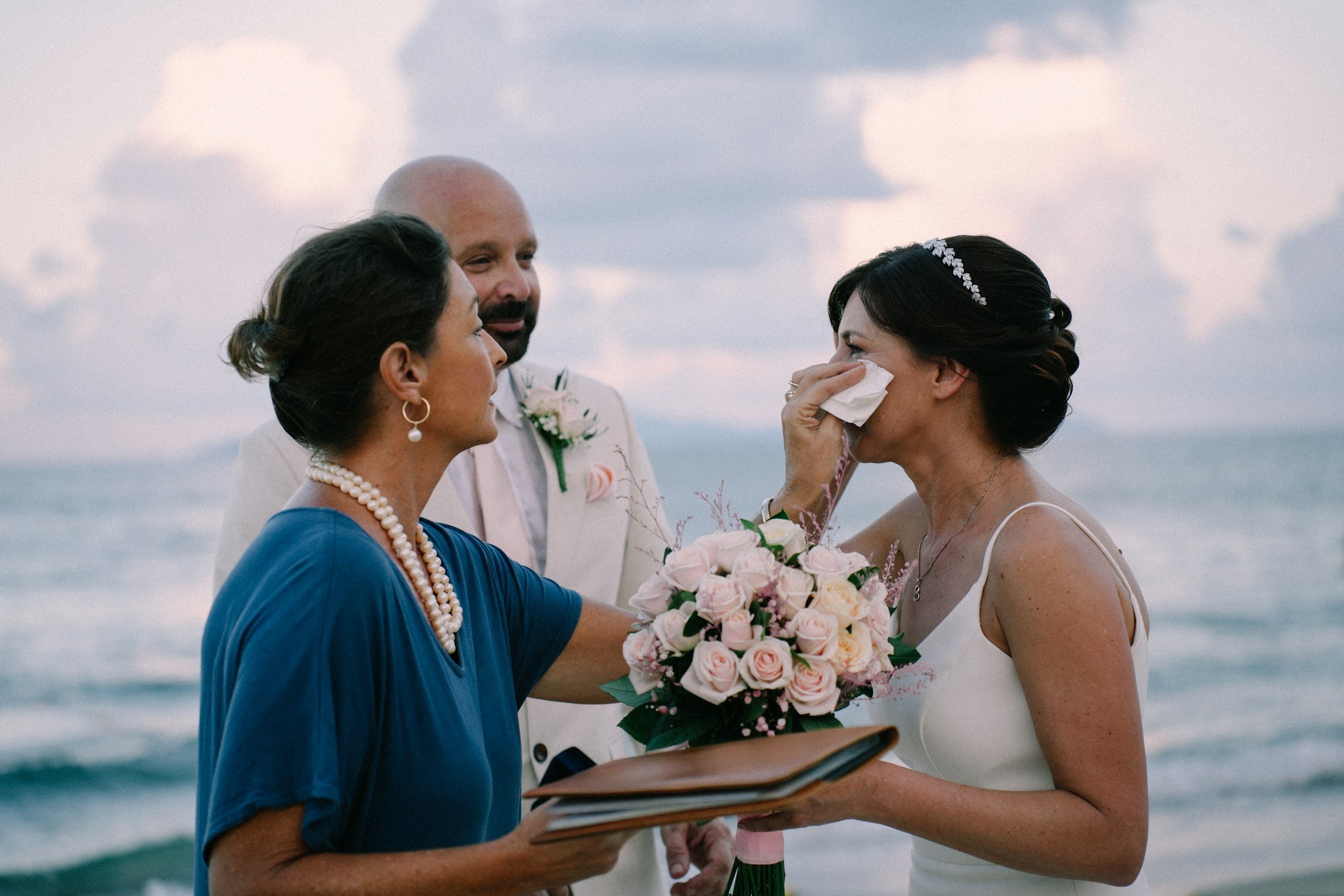 A bride with dark hair in an updo, wearing a white dress and a pearl headband, is crying and holding a bouquet of pink roses while wiping her tears with a tissue. An older woman in a blue dress with pearl jewelry is comforting her, and a man in a light suit is standing behind them at the beach during sunset.
