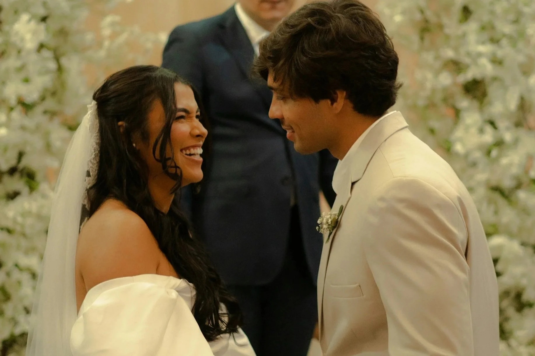 A couple dressed in wedding attire smiling and looking at each other during their wedding ceremony, with a person standing behind them and a background of white flowers.