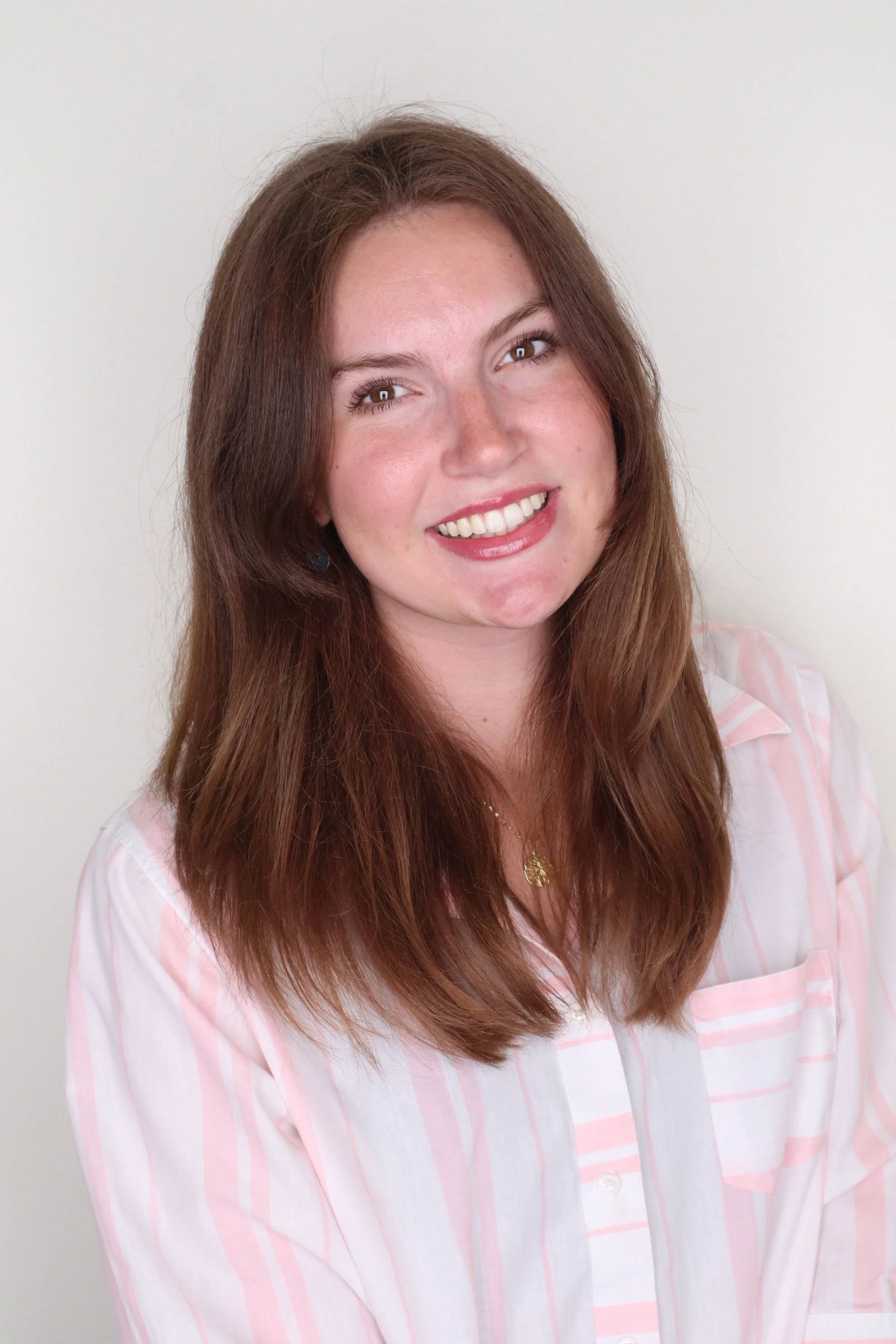 A young woman with long, reddish-brown hair smiling at the camera, wearing a light pink and white striped shirt and a gold necklace, with a plain off-white background.