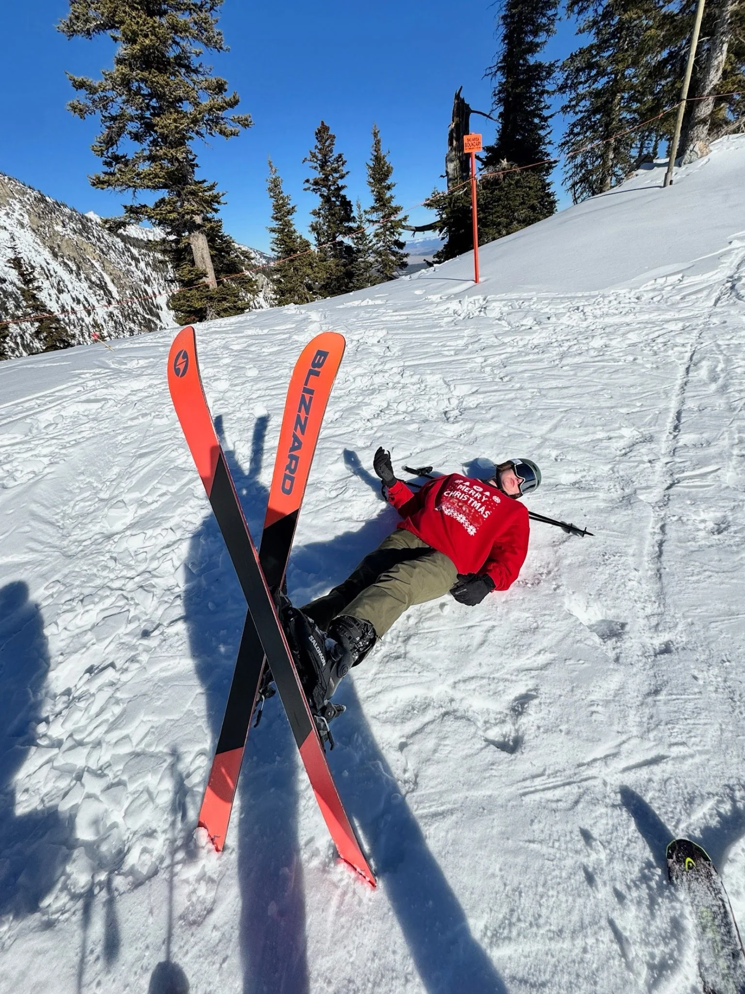 A person lying on snow next to red and black skis with the word 'BLIZZARD' on them, wearing a red Christmas sweater, black gloves, helmet, and sunglasses, on a snowy mountain slope with pine trees, a clear blue sky, and ski trail signs in the background.
