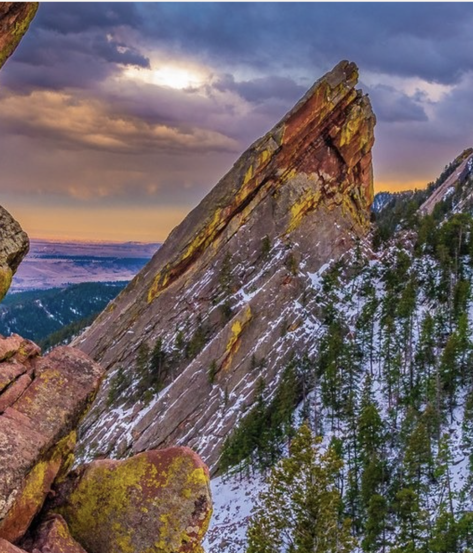 Large pointed mountain with snow and green trees at the base, under a colorful sky during sunset or sunrise.