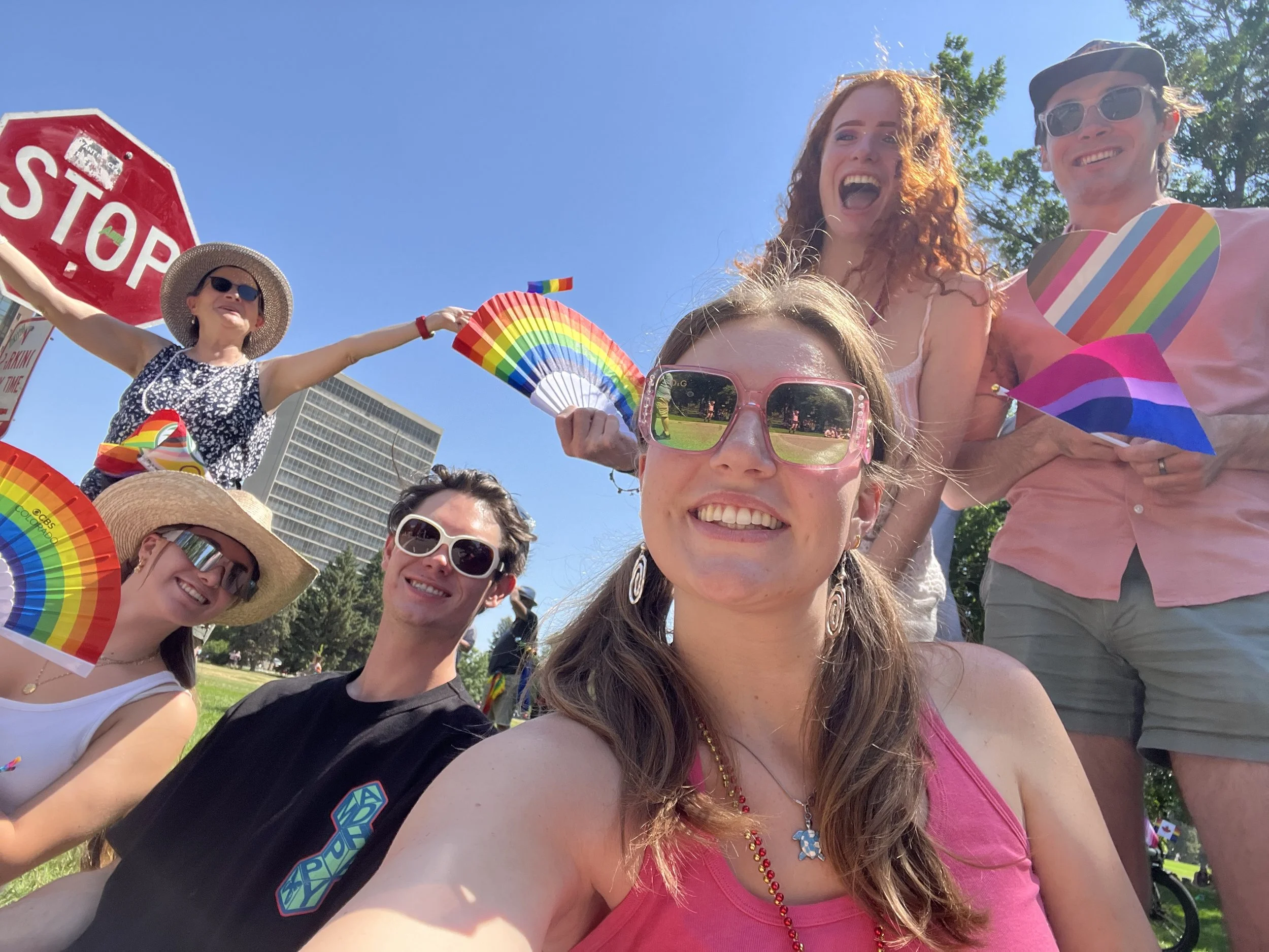 Group of people celebrating outdoors with rainbow-themed accessories, smiling and posing for a selfie on a sunny day.