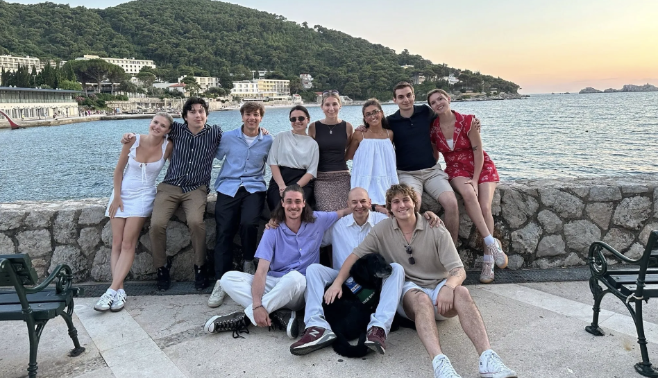 Group of ten friends posing by the water at sunset, with hills and buildings in the background.