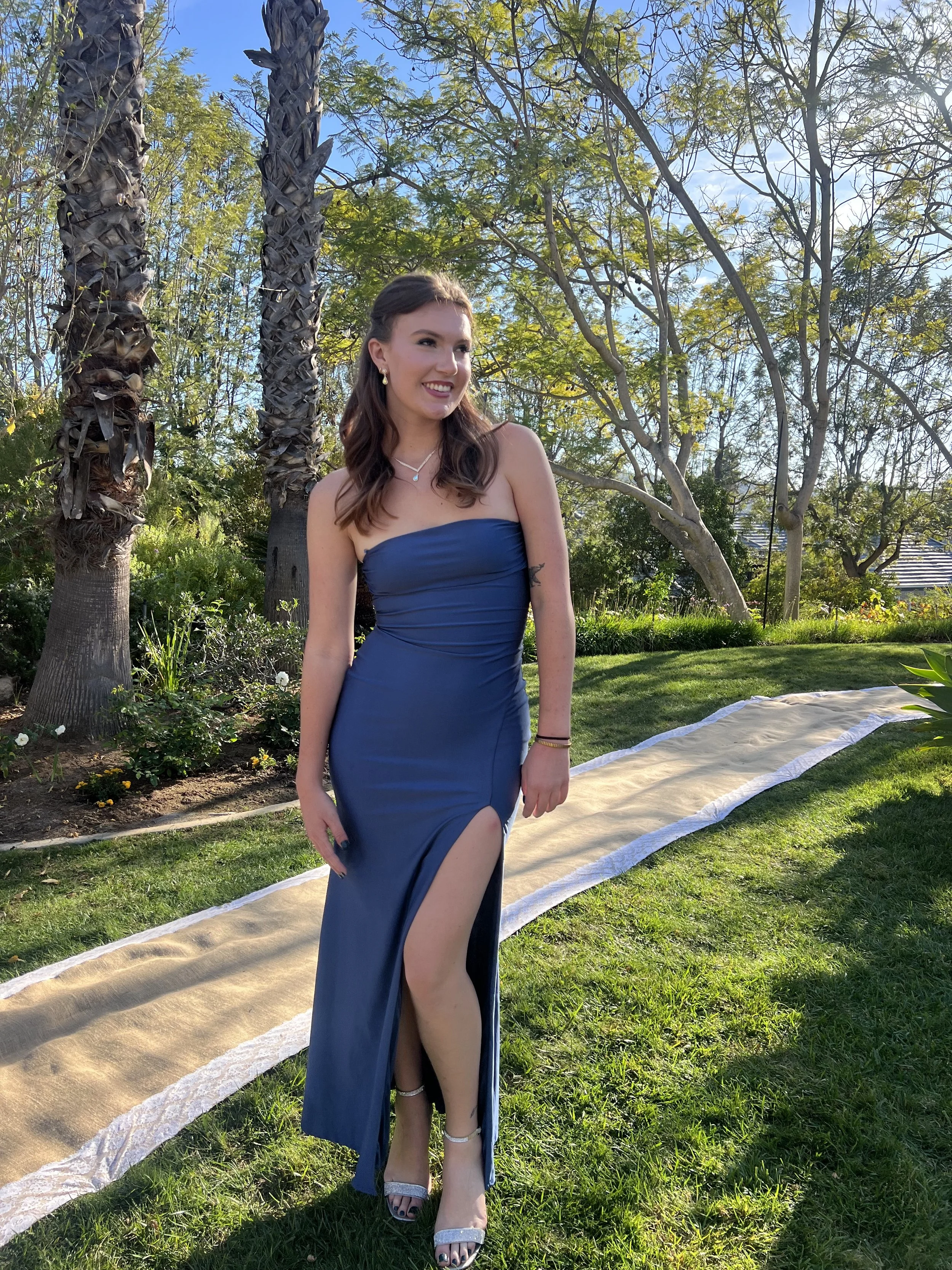 A young woman in a strapless blue gown with a thigh-high slit, standing on a grassy lawn during daytime, smiling and posing outdoors near palm trees and other greenery.