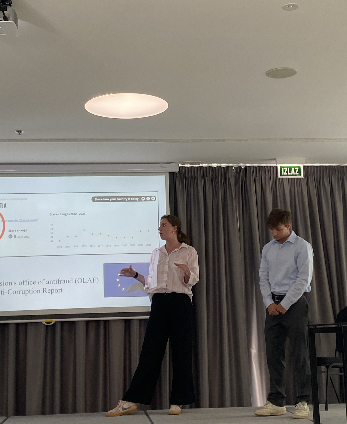 Two people giving a presentation in a conference room, with a large screen displaying a chart about European Union countries.