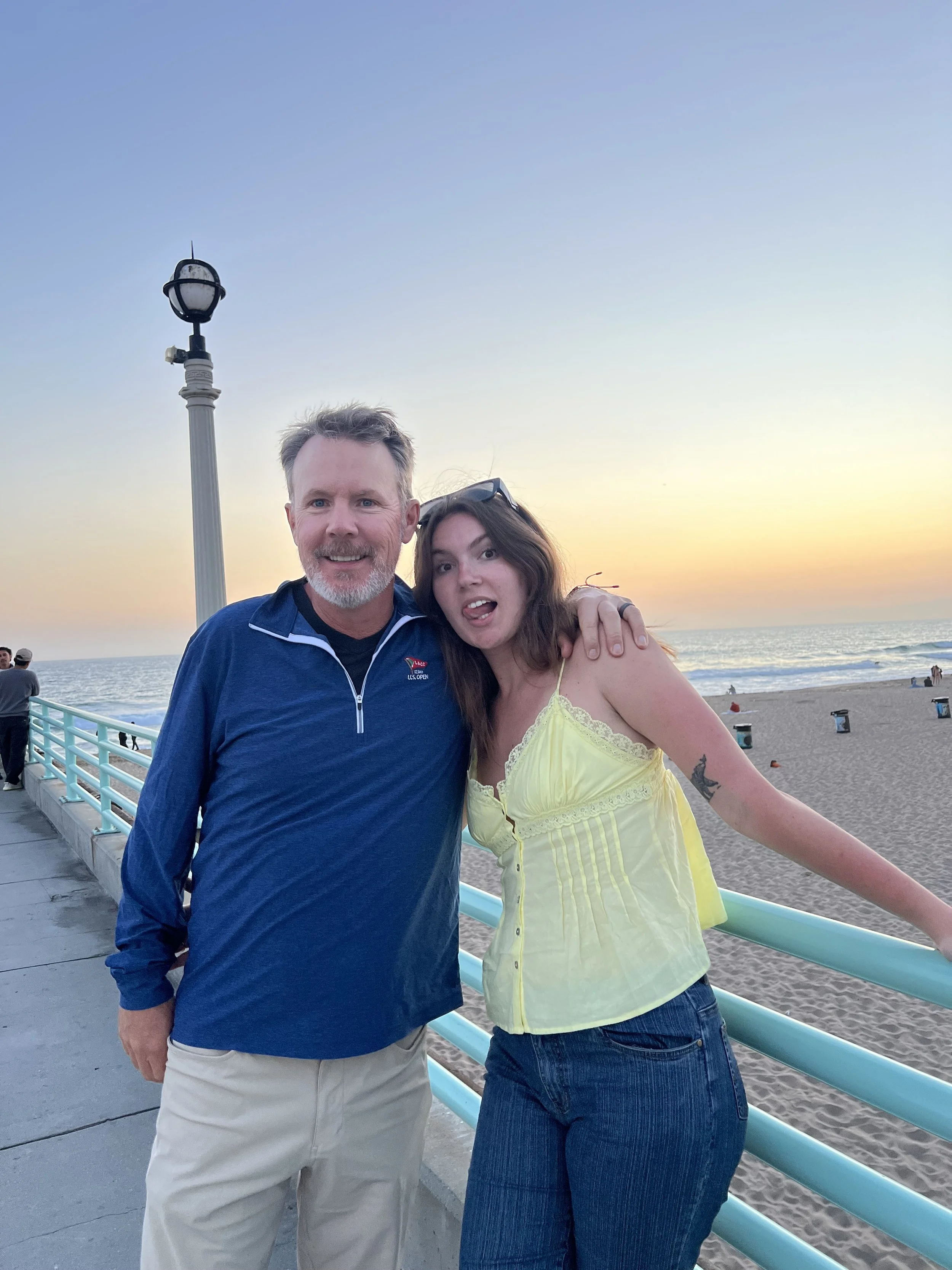 A man and woman posing together on a beachside pier during sunset. The man is wearing a blue jacket and khaki pants, and the woman is wearing a yellow top and jeans. They are smiling and appear to be enjoying the moment.