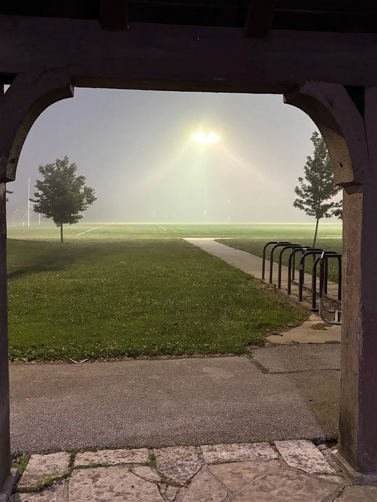 A foggy sports field visible through a stone archway at night, illuminated by bright stadium lights, with a pathway and bike rack in the foreground.
