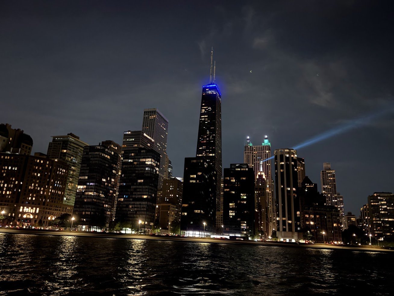 Nighttime city skyline with tall skyscrapers, including a distinctive tower with blue lights on top, reflecting off the water in the foreground.