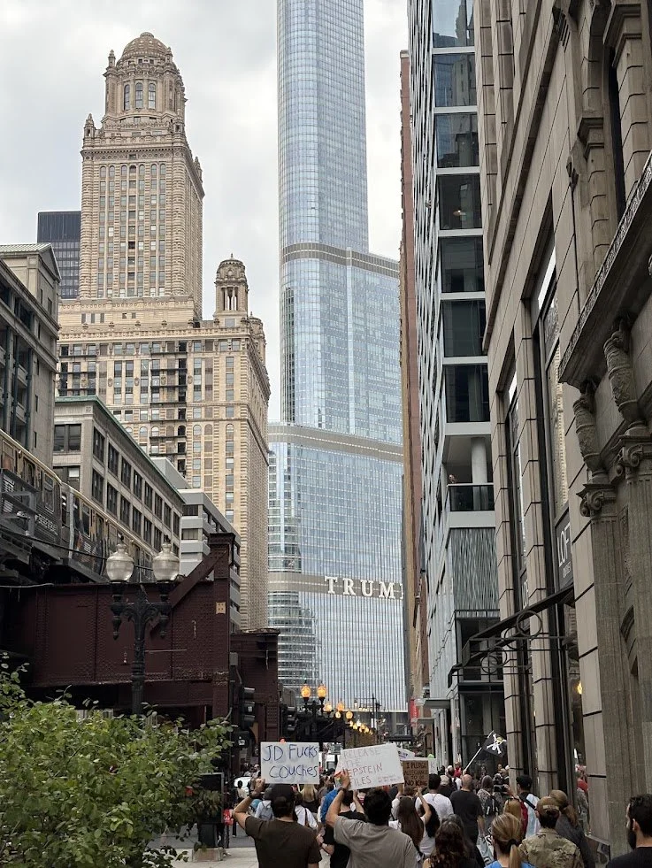 Protesters gathering on a city street in front of a tall building with a sign saying "TRUMP." The crowd holds various signs, and the surrounding cityscape includes historic and modern skyscrapers.