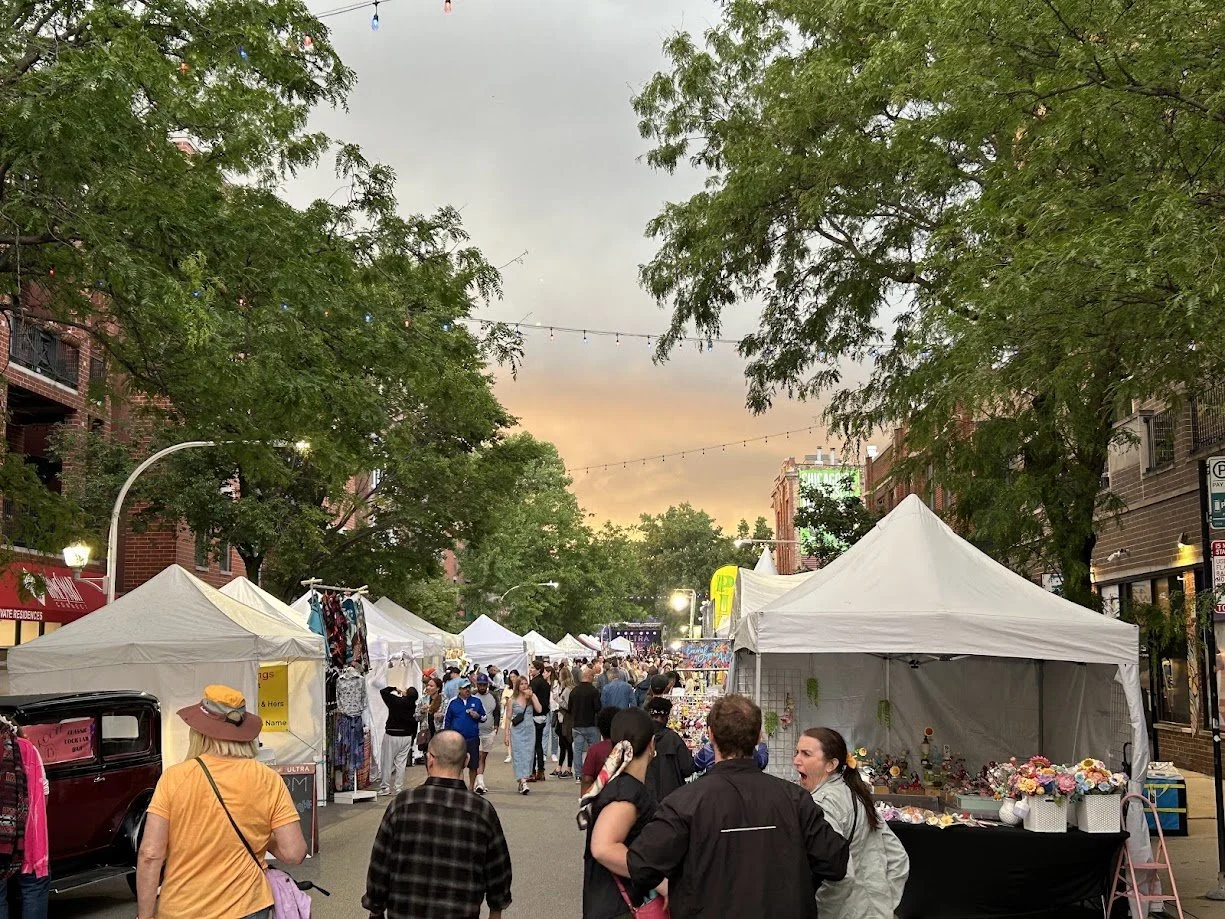 Outdoor street fair with white tents, people browsing, and vendors. Trees line the street, and a sunset sky is visible.