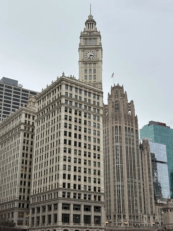 A tall historic clock tower building in a city with other skyscrapers around it, overcast sky.