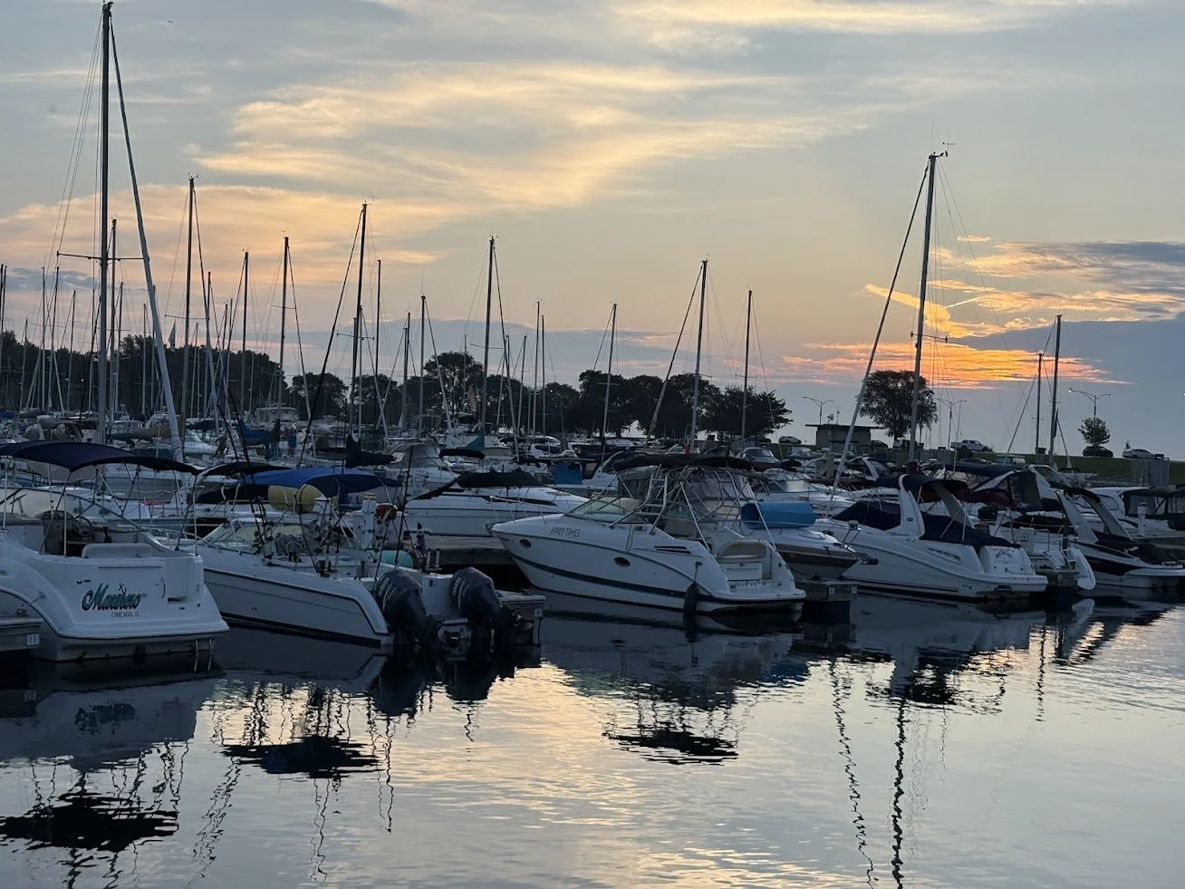 A marina with multiple docked boats and yachts at sunset, calm water reflecting the vessels and a partly cloudy sky with the sun setting behind trees.