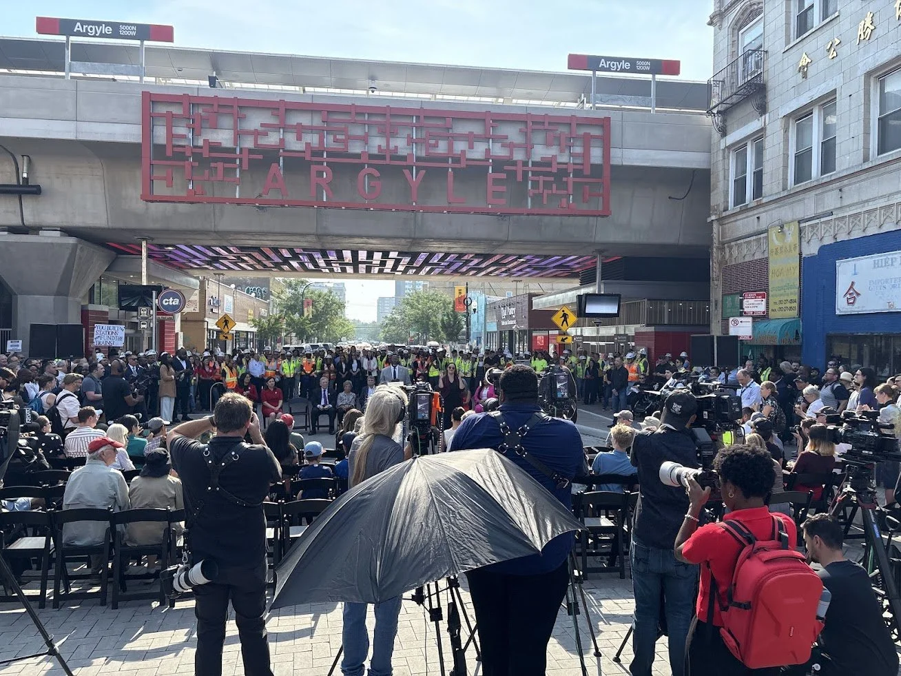 A crowd gathered for a public event or press conference outside a building with a large sign reading 'Argyle' and 'Argyle' street signs, with photographers and cameras capturing the scene.